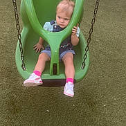 Kayliah is registered to the contest to win money with this photo: toddler, child, swing, playground, green_swing, adaptive_swing, grass, outdoor, fence, parked_cars, shoes, pink_socks, denim_overalls, shorts, hair_tie, daylight, person, seat, leisure, play