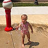 Kayliah joined the competition — help win amazing prizes! toddler, child, water_fountain, splash_pad, swimsuit, playing, water_splash, outdoor, sunlight, shadow, concrete, baseball_post, playground, person, grass, park, summer, happy, fun, daytime