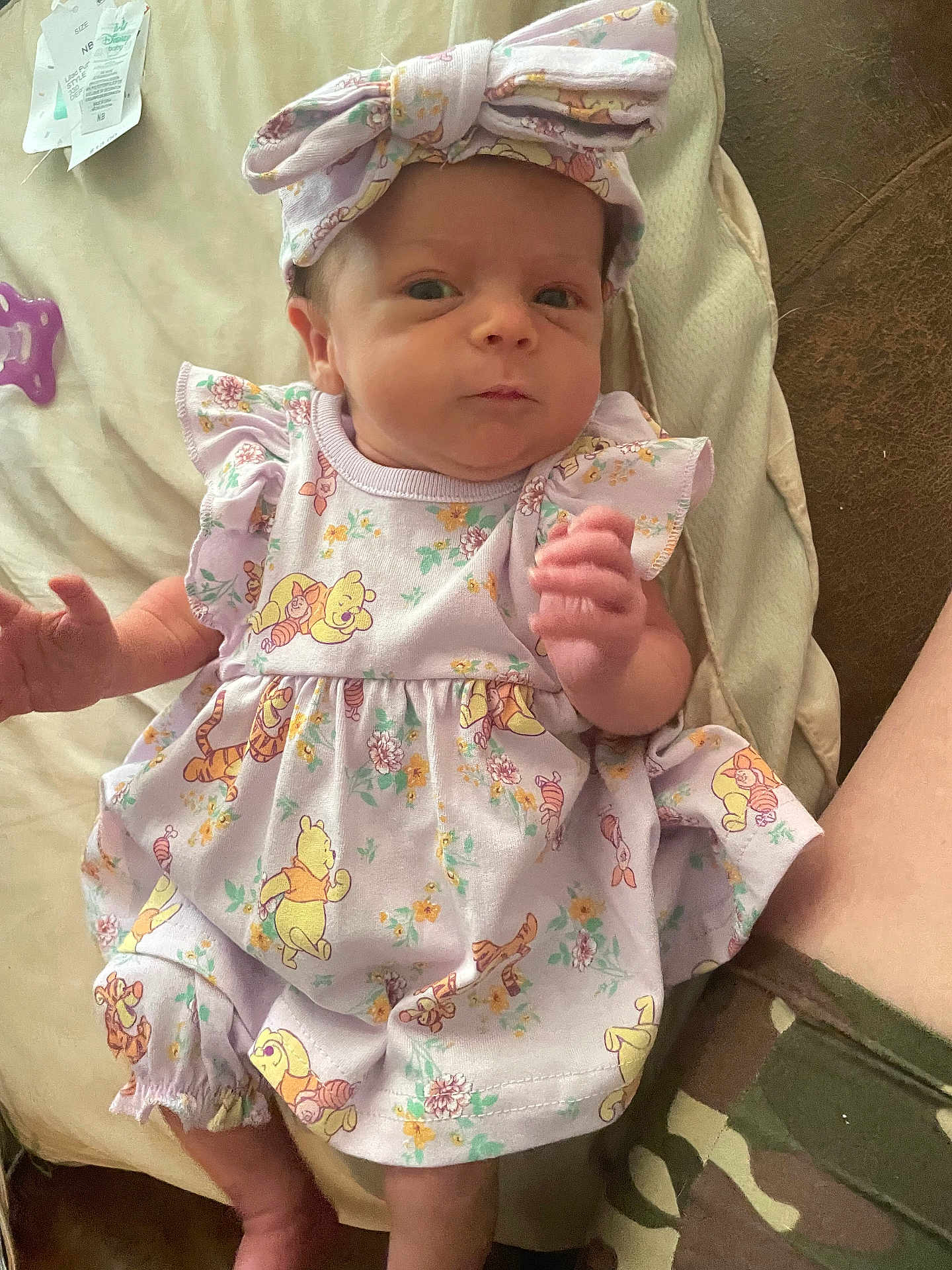 Mazzie is registered to the contest to win money with this photo: baby, infant, child, purple_outfit, headband, winnie_the_pooh, patterned_clothing, lying_down, soft_surface, hand, face, person, legs, shorts, camouflage, curious_expression, indoors, closeup, cute, newborn