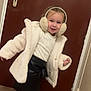 child, girl, earmuffs, white_jacket, black_skirt, tights, indoor, wooden_door, flooring, playful, smiling, portrait, fashion, winter_clothing, cute, happy, person, standing, cozy, home