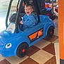 child, toddler, toy_car, blue_car, smiling, indoor, shopping_mall, ride_on_toy, floor_tiles, happy, person, play, kid, vehicle, fun, casual_clothing, hoodie, plastic, wheels, entertainment