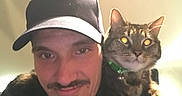 Simba is registered to the contest to win money with this photo: man, cat, tabby_cat, indoor, portrait, cap, smile, pet, shoulder, green_collar, facial_hair, casual_clothing, closeup, human, animal, friendship, domestic, eyes, feline, mammal