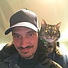 animal, cap, casual_clothing, cat, closeup, domestic, eyes, facial_hair, feline, friendship, green_collar, human, indoor, mammal, man, pet, portrait, shoulder, smile, tabby_cat