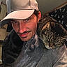 Simba joined the competition — help win amazing prizes! man, cat, indoor, cap, gray_shirt, tabby_cat, smile, facial_hair, pet, shoulder, relaxed, cozy, affectionate, animal, human, portrait, casual, furniture, curtain, home