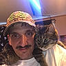 Simba is registered to the contest to win money with this photo: man, cat, tabby_cat, indoor, portrait, hat, camo_hat, white_shirt, mustache, pet, animal, shoulder, face, feline, looking_at_camera, cozy, human, light_streak, background, closeup