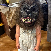 Brianna is registered to the contest to win money with this photo: child, mask, werewolf_mask, costume, indoor, wooden_floor, romper, pink_sandals, furniture, couch, blanket, toy, scary, face_covering, hair, person, standing, room, home, playful