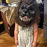 child, mask, werewolf_mask, costume, indoor, wooden_floor, romper, pink_sandals, furniture, couch, blanket, toy, scary, face_covering, hair, person, standing, room, home, playful