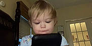 Brianna is registered to the contest to win money with this photo: bed, blinds, casual_clothing, child, curtain, device, door, face, focus, furniture, hair, hand, indoor, light, person, phone, room, toddler, window, young_child