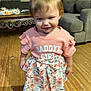 toddler, child, girl, pink_shirt, floral_skirt, bow, barefoot, wooden_floor, living_room, sofa, table, smile, short_hair, indoors, cute, person, clothing, floor, home, portrait