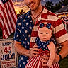 baby, man, patriotic_clothing, fireworks, american_flag, outdoor, celebration, holiday, independence_day, smiling, holding, headband, bow, dress, shirt, evening, garden, sign, flowers, festive