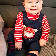 Lysandre participe au concours pour gagner de l'argent avec cette photo : baby, child, smiling, sitting, striped_clothing, overalls, bear_design, couch, blanket, indoor, happy, face, hands, feet, socks, striped_socks, furniture, young_child, portrait, casual