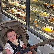 Roman is registered to the contest to win money with this photo: baby, bakery, cakes, child, clothing, curious, desserts, face, food_display, indoor, infant, pastries, person, retail, seat, shopping, socks, store, stroller, window
