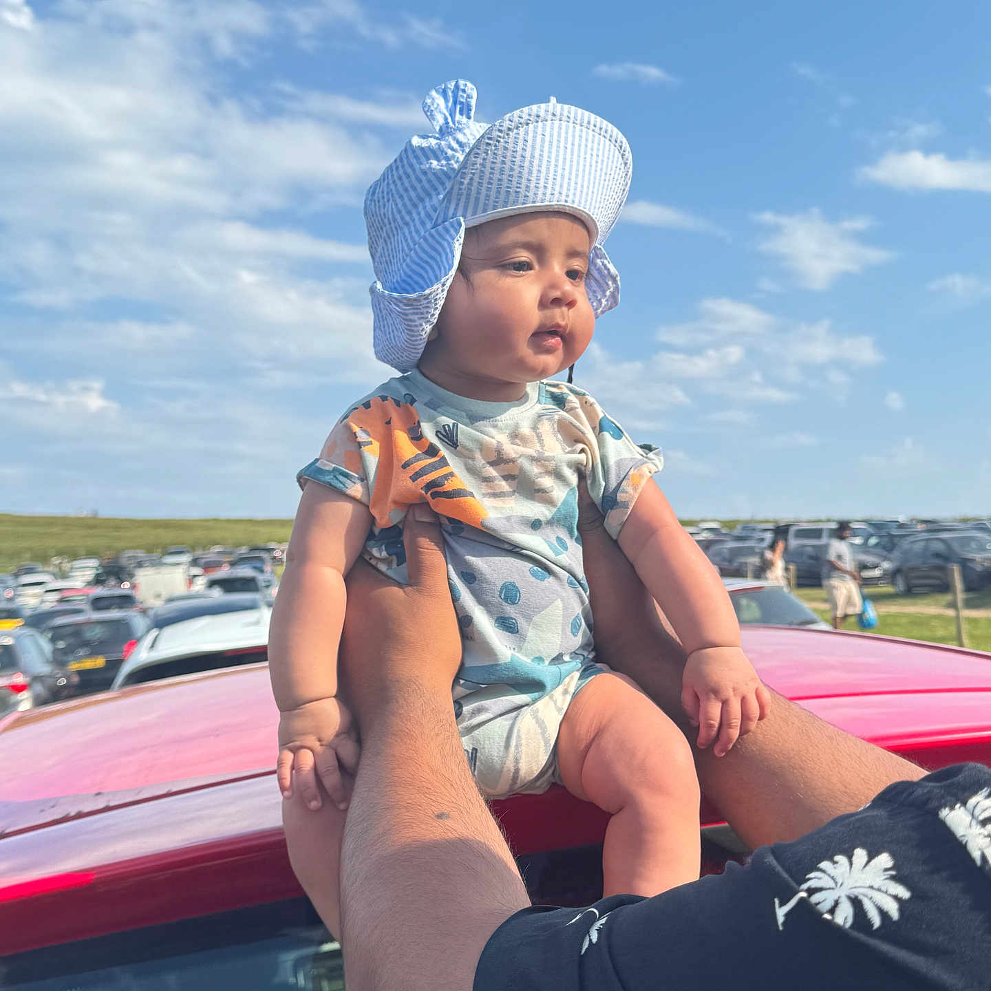 Rudra is registered to the contest to win money with this photo: adult_hands, baby, car, casual_clothing, child, clouds, cute, daytime, family, hat, holding, infant, nature, outdoor, parking_lot, people, person, sky, summer, sun_hat