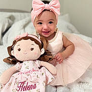 Jocelyn joined the competition — help win amazing prizes! baby, child, doll, pink_dress, headband, bow, plush_toy, smile, jewelry, pearls, soft_toy, bed, blanket, indoor, cute, happy, person, portrait, toddler, sitting