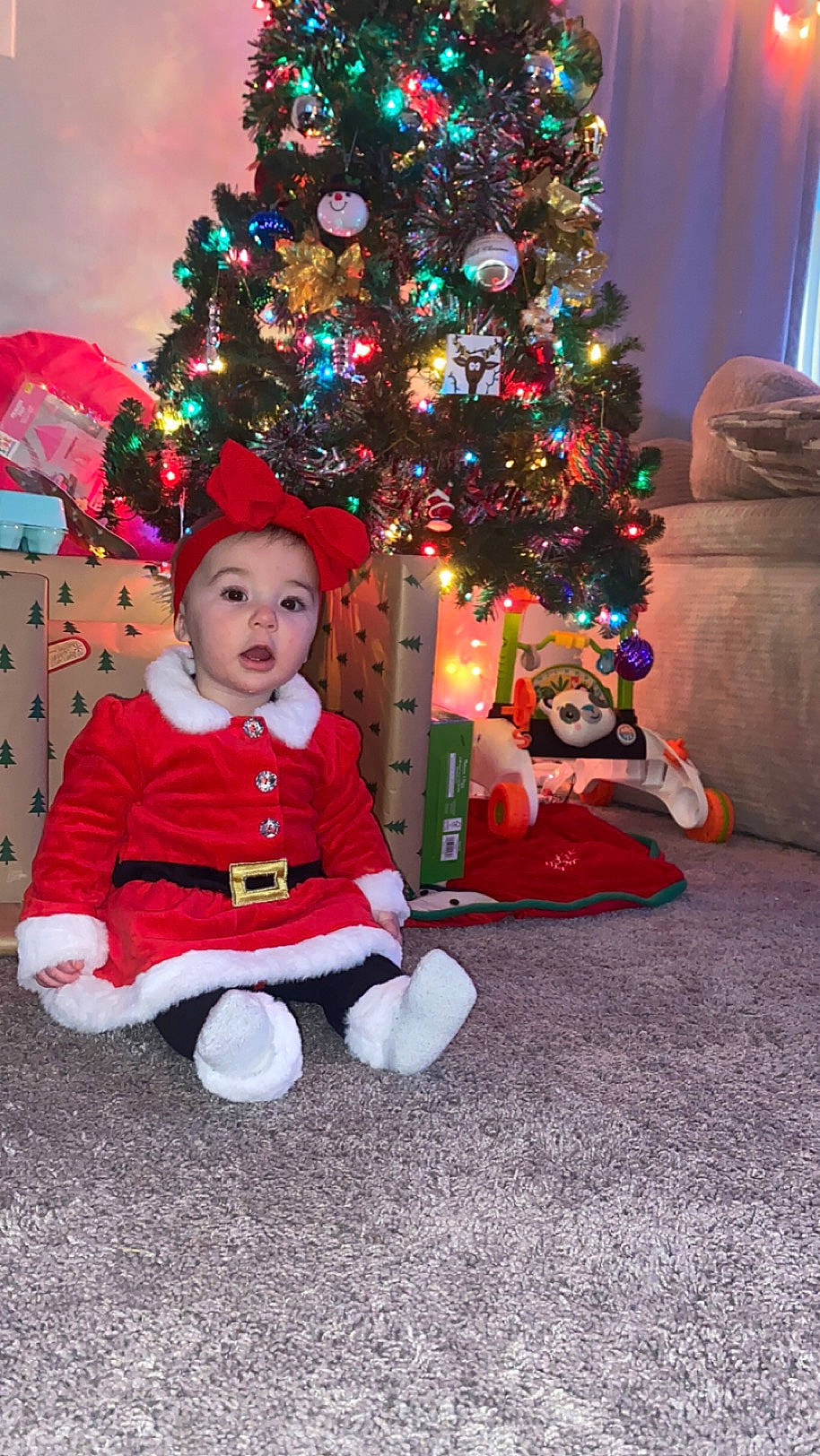 Violet is registered to the contest to win money with this photo: baby_toddler_clothing, christmas, christmas_decoration, christmas_eve, christmas_ornament, christmas_tree, conifer, event, facial_expression, flooring, fun, happy, headwear, holiday, holiday_ornament, lap, ornament, person, plant, surprise