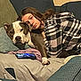 blanket, brown_dog, casual, checkered_shirt, comfort, companion, couch, dog, furniture, home, indoor, living_room, paw, person, phone, plaid_clothing, relaxing, resting, snuggling, white_dog