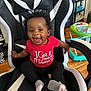 baby, child, smile, happy, sitting, red_shirt, black_pants, gray_socks, gaming_chair, curly_hair, earrings, indoor, floor, toys, walker, furniture, person, expression, home, wooden_floor