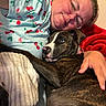 dog, dogs, brindle_dog, puppy, person, human, glasses, face, cuddling, hugging, indoors, bed, blanket, red_blanket, pajamas, cherry_pattern, pet, portrait, cozy, home