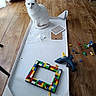cat, white_cat, heterochromia, cardboard_box, lego, lego_bricks, glue_gun, wooden_floor, indoor, pet, animal, floor, craft, project, toys, scattered, sitting, curious, domestic, home