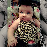Salem is registered to the contest to win money with this photo: baby, infant, child, portrait, sitting, stroller, leopard_print, animal_print, dress, headband, dark_hair, wide_eyes, face, hands, indoor, furniture, blanket, cushion, colorful, photograph