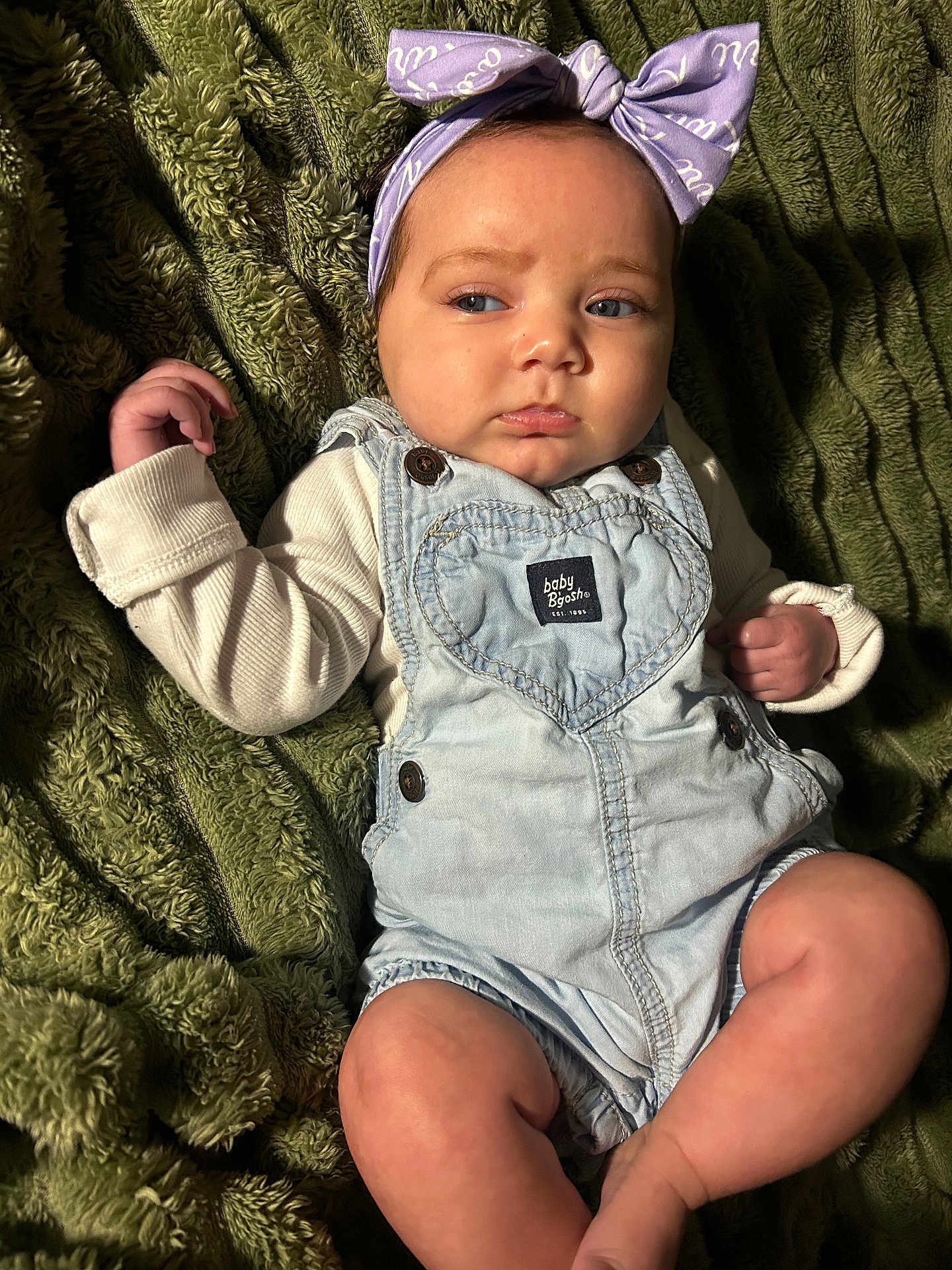 Kari joined the competition — help win amazing prizes! baby, child, infant, headband, bow, denim_overalls, clothing, blanket, green_background, skin, face, legs, hand, portrait, indoor, cute, expression, young, person, sitting