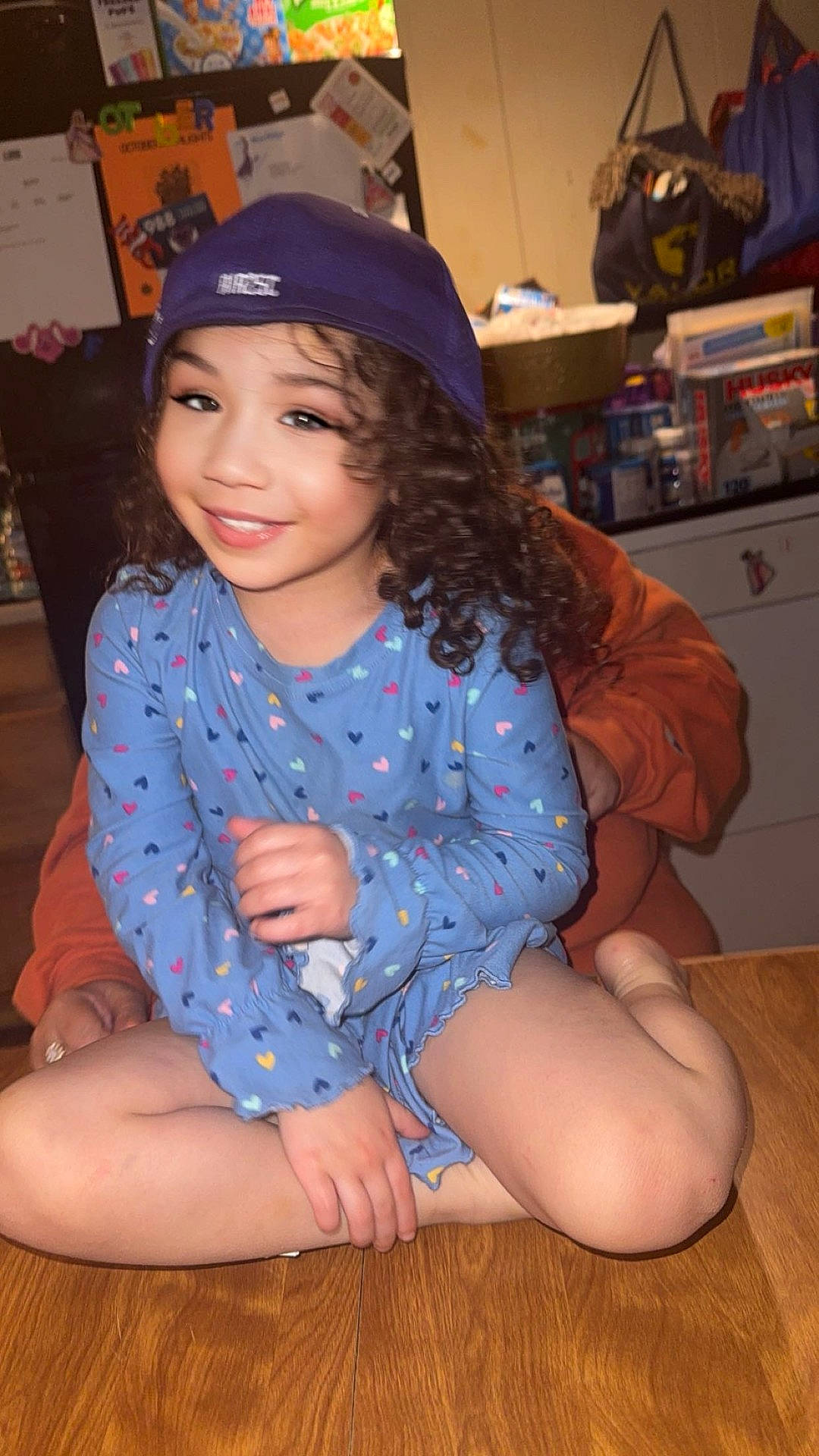 Mya is registered to the contest to win money with this photo: adult, background_clutter, blue_dress, cabinet, casual_clothing, child, cross_legged, curly_hair, family, happy, indoor, kitchen, notes, orange_shirt, person, portrait, purple_cap, refrigerator, smiling, wooden_table