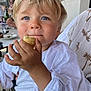 blonde_hair, blue_eyes, casual, child, closeup, cute, dining, face, food, hand, happy, indoor, light, pastry, person, portrait, table, toddler, white_shirt, young_child