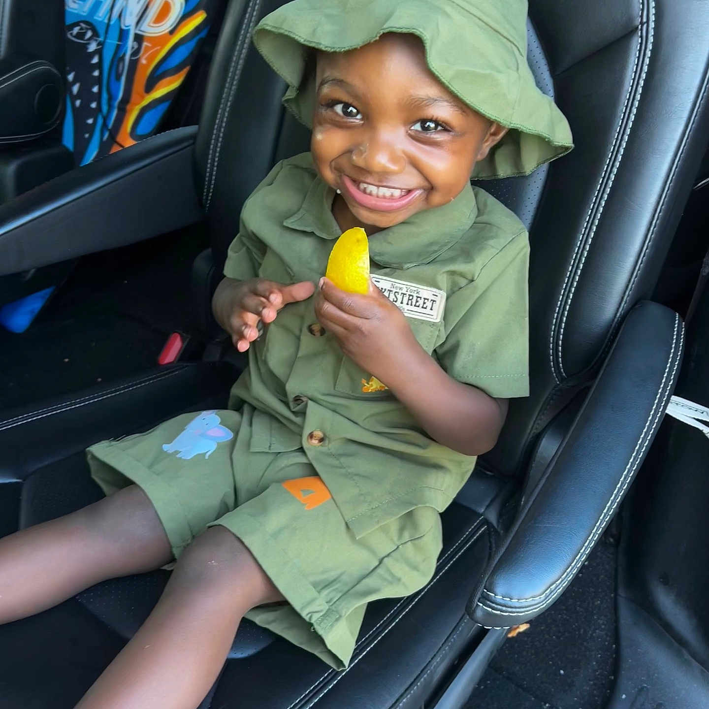 Ezra is registered to the contest to win money with this photo: child, toddler, smile, green_clothing, hat, car_seat, toy, yellow_object, happy, person, seat, interior, car, footwear, socks, cute, portrait, kid, seatbelt, vehicle