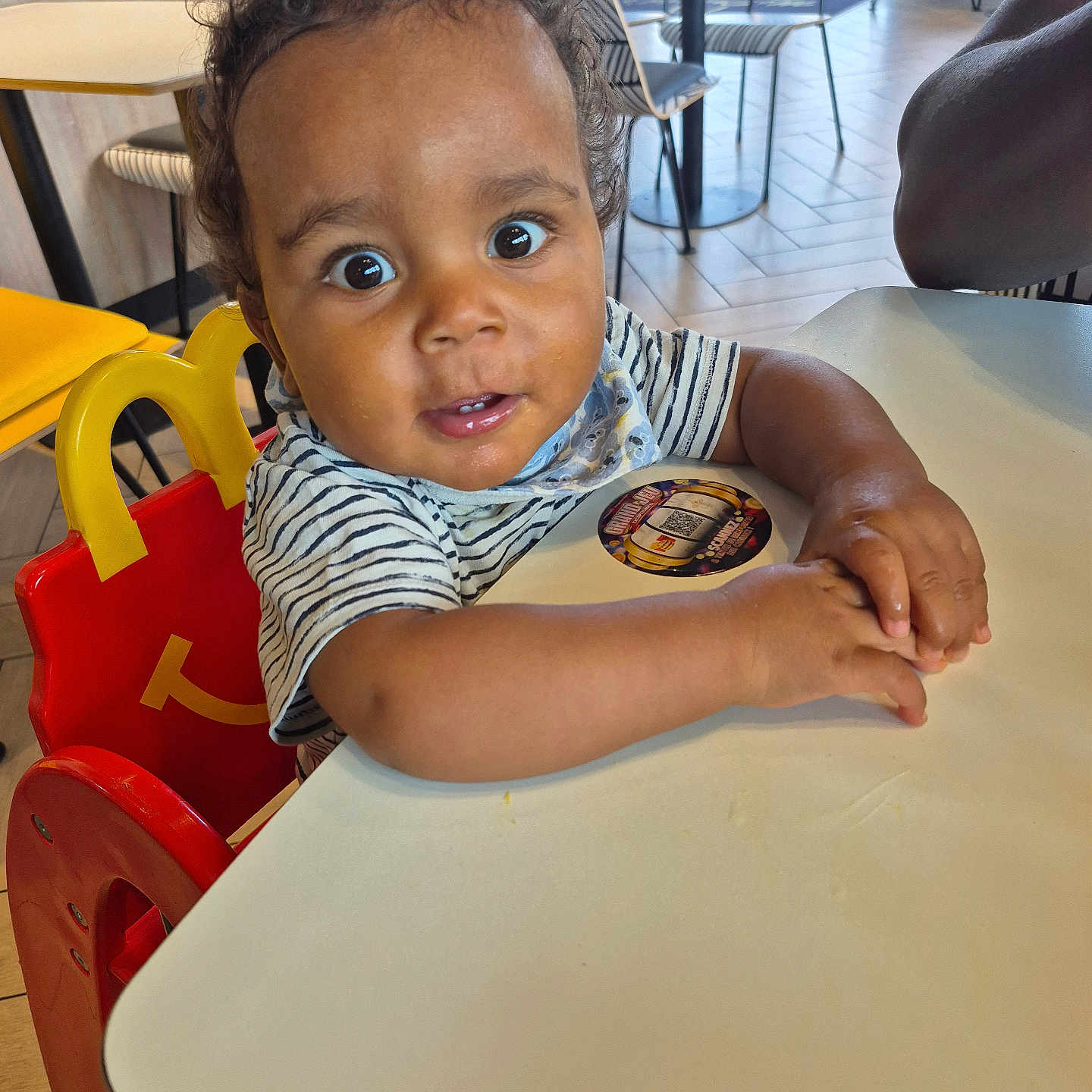 Adonys participe au concours pour gagner de l'argent avec cette photo : background, casual_clothing, child, curious, curly_hair, expression, face, floor, furniture, hands, high_chair, indoor, light, person, restaurant, seat, striped_shirt, table, toddler, wide_eyes