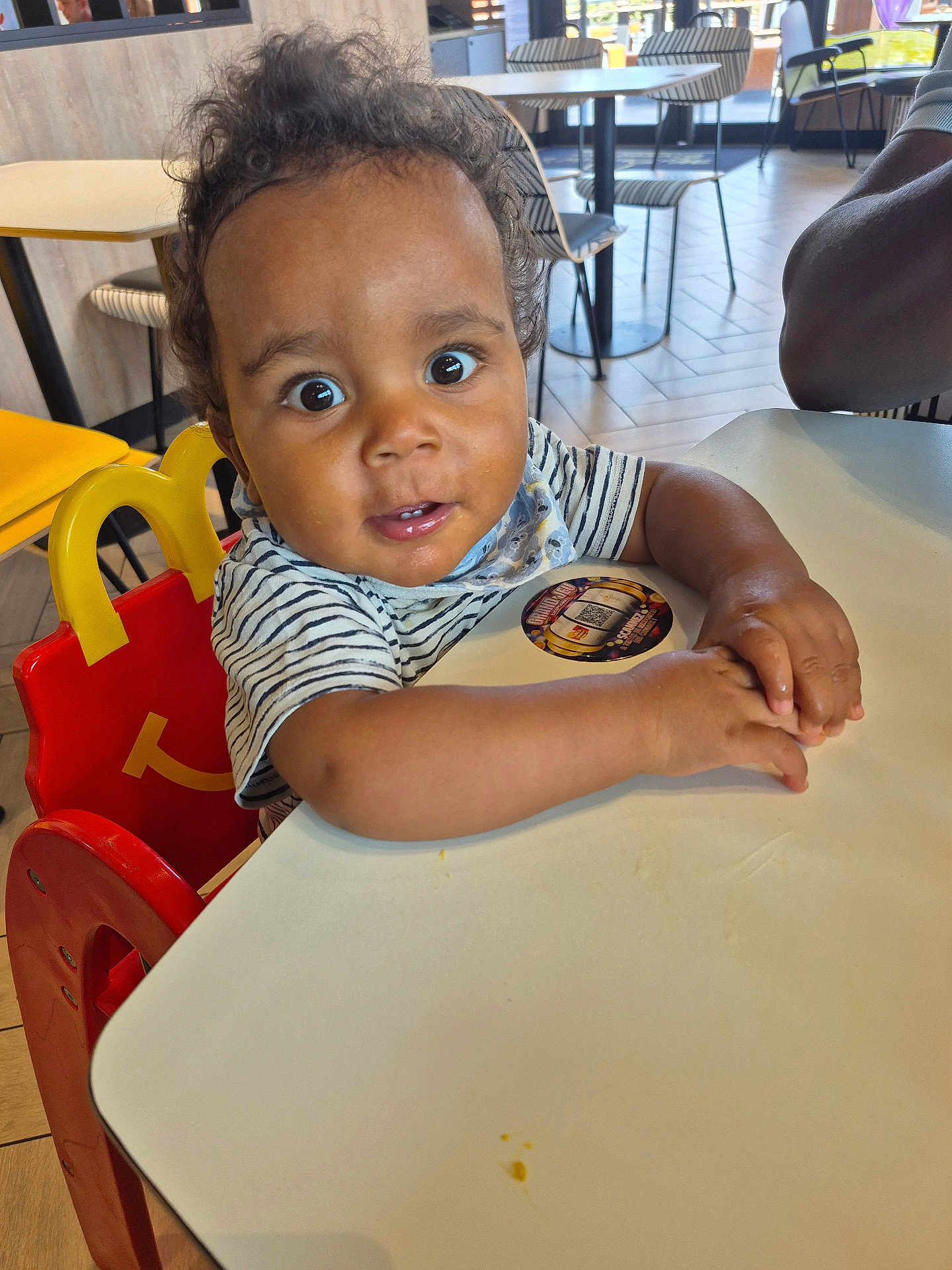 Adonys participe au concours pour gagner de l'argent avec cette photo : toddler, child, high_chair, restaurant, table, curly_hair, striped_shirt, indoor, wide_eyes, hands, furniture, floor, person, face, seat, expression, casual_clothing, background, light, curious