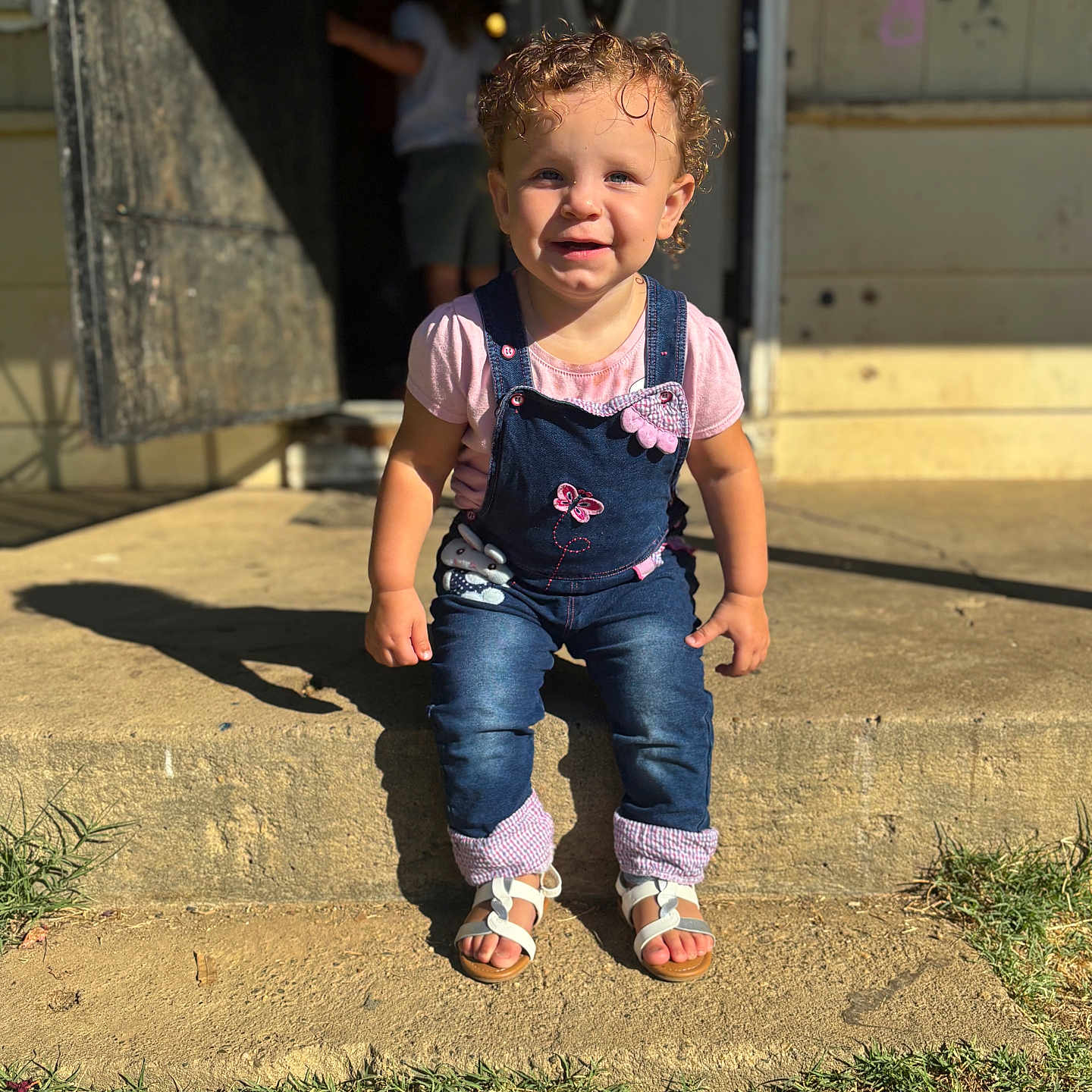 Amiliyah is registered to the contest to win money with this photo: child, concrete_step, curly_hair, cute, daylight, denim_overalls, face, front_yard, grass, happy, outdoor, person, pink_shirt, portrait, sandals, shadow, smiling, sunlight, toddler, young_child