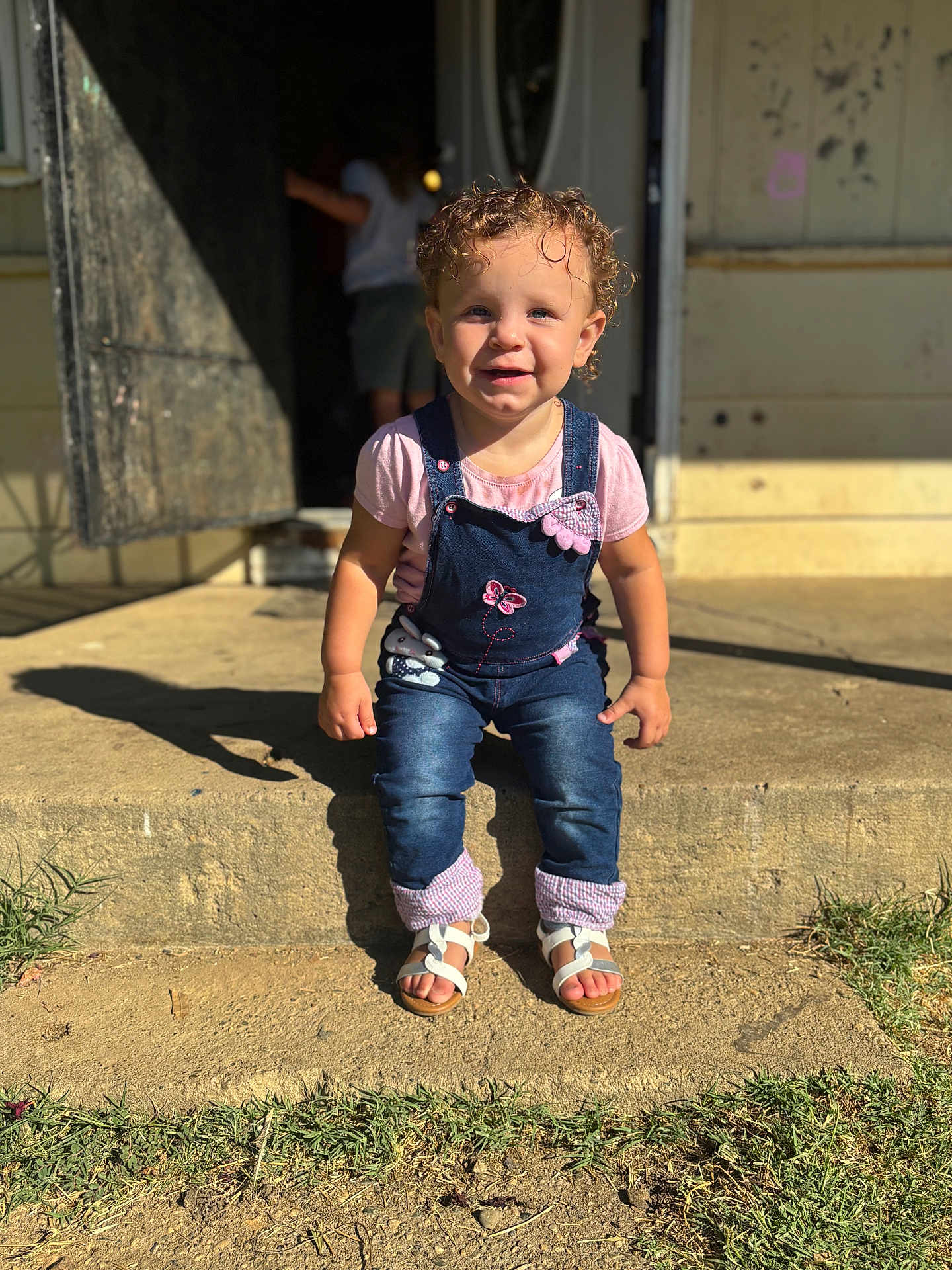 Amiliyah is registered to the contest to win money with this photo: toddler, child, curly_hair, denim_overalls, pink_shirt, sandals, outdoor, sunlight, shadow, concrete_step, grass, smiling, face, person, front_yard, daylight, cute, happy, young_child, portrait