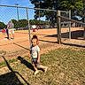 Mateo is registered to the contest to win money with this photo: child, baseball_field, fence, grass, tree, person, sunlight, shadow, sports, outdoor, blue_sky, shorts, tshirt, young_child, daytime, recreation, people, playing, hat, casual_clothing