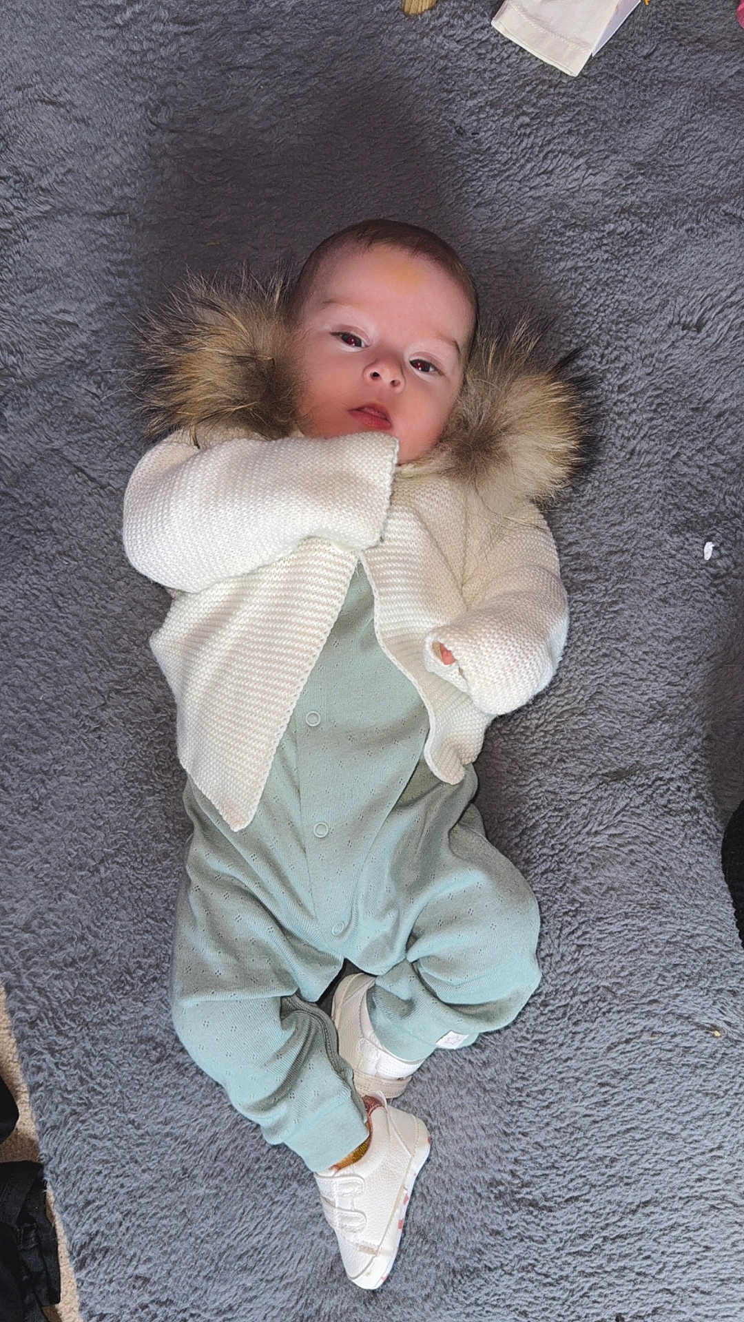 Isabel joined the competition — help win amazing prizes! baby, infant, blanket, clothing, sweater, fur, footed_pajamas, shoes, lying_down, face, head, person, portrait, soft_texture, gray_background, cute, young_child, comfortable, warm_clothing, indoors