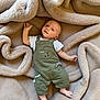 baby, infant, child, overalls, green_clothing, dinosaur_design, blanket, soft_texture, beige, laying_down, happy_expression, cute, small_hand, small_foot, indoor, portrait, smiling, face, head, cozy