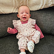 Emerie is registered to the contest to win money with this photo: baby, infant, smiling, sitting, couch, blanket, knitted, clothing, footwear, white_shoes, happy, portrait, indoor, cute, child, person, face, apparel, fabric, cozy