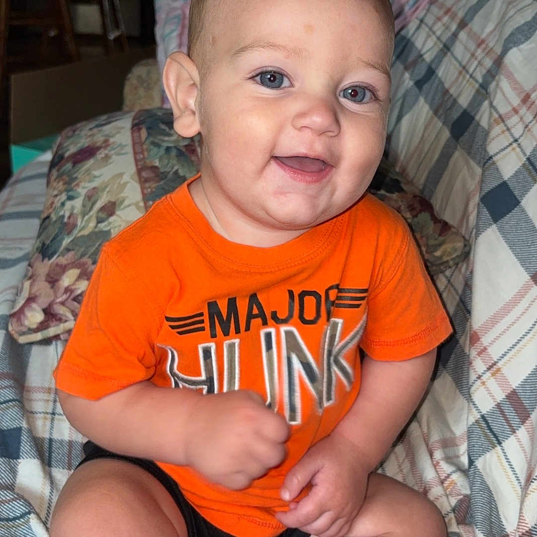 Jasper joined the competition — help win amazing prizes! baby, child, infant, smiling, happy, portrait, sitting, orange_shirt, plaid_couch, blanket, indoors, home, face, eyes, short_hair, barefoot, casual_clothing, playful, photography, couch