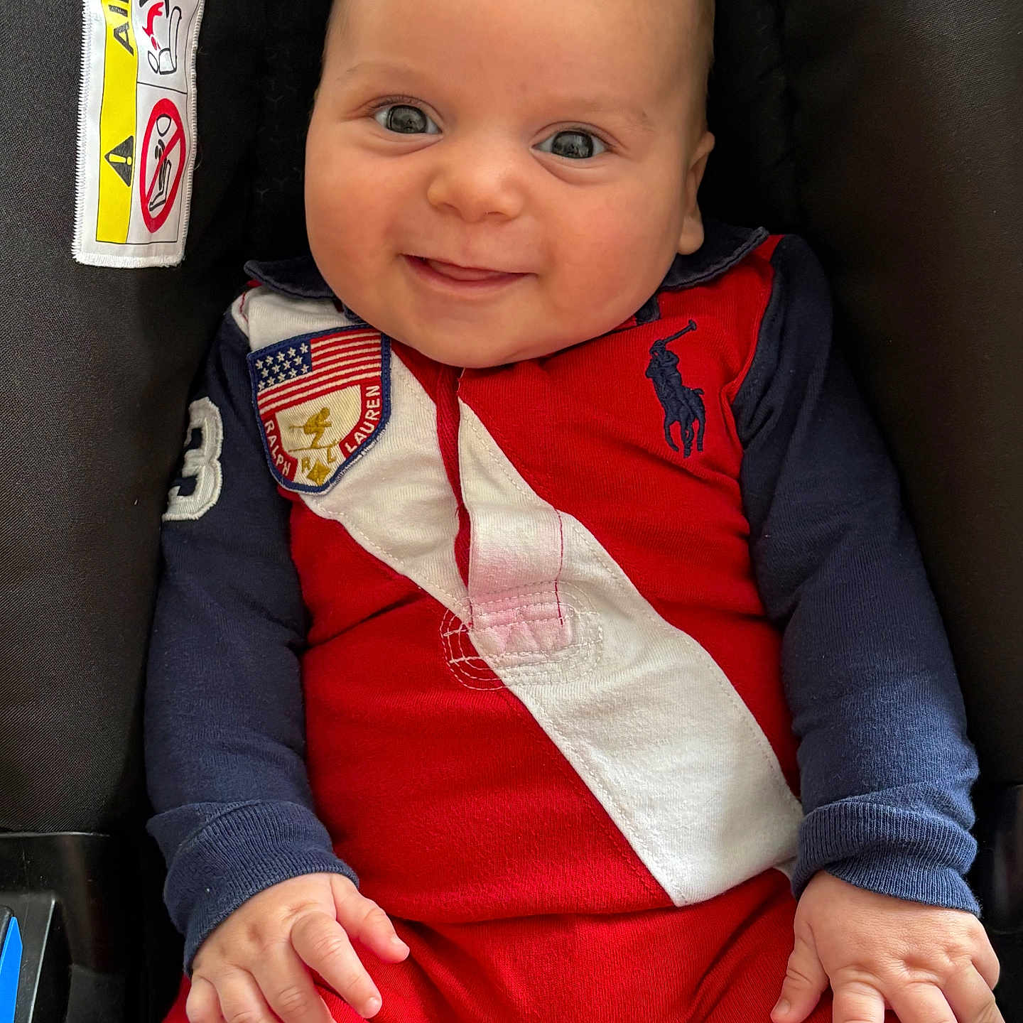 Gabriel-Moussa participe au concours pour gagner de l'argent avec cette photo : airbag, baby, car_seat, child, closeup, clothing, cute, face, hands, happy, indoor, infant, navy_sleeves, person, portrait, red_outfit, seated, smiling, warning_label, young