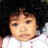 child, toddler, portrait, close_up, curly_hair, brown_skin, cute, eyes, face, expression, white_clothing, pink_embroidery, indoor, baby, young_child, person, innocence, adorable, headshot, looking