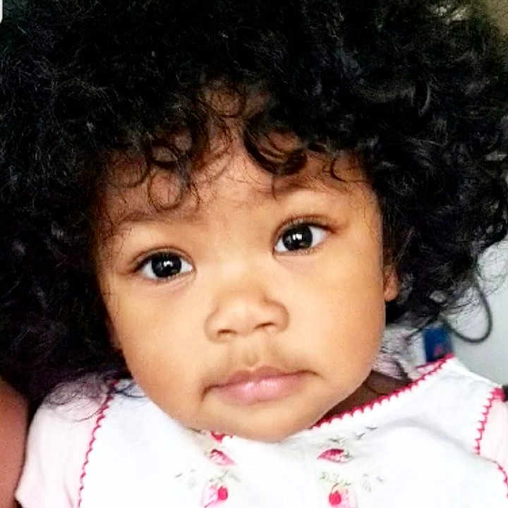 Monica joined the competition — help win amazing prizes! child, toddler, portrait, close_up, curly_hair, brown_skin, cute, eyes, face, expression, white_clothing, pink_embroidery, indoor, baby, young_child, person, innocence, adorable, headshot, looking