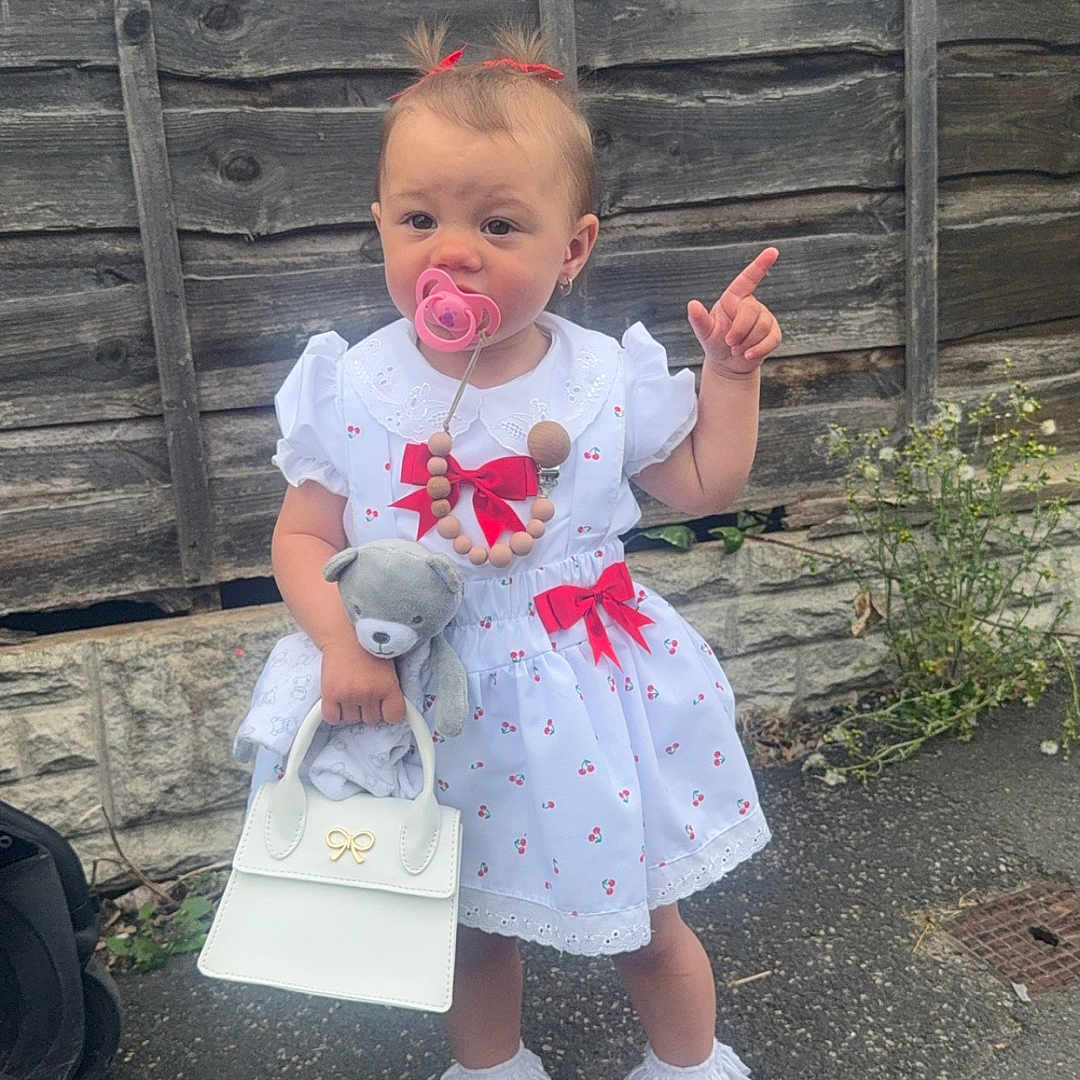 Vienna-Raine joined the competition — help win amazing prizes! accessory, child, cute, fashion, frilly_socks, hair_ribbons, handbag, outdoor, pacifier, pavement, person, red_bows, standing, stone_wall, teddy_bear, toddler, toy, white_dress, white_shoes, wooden_fence