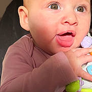 Myla is registered to the contest to win money with this photo: baby, child, infant, toy, hand, face, eyes, mouth, pink_clothing, cute, closeup, skin, expression, holding, person, small_hand, indoor, adorable, young_child, happy