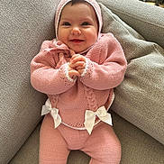 Lyra a rejoint le concours — aidez-le/la à gagner de superbes lots ! baby, smile, pink_clothing, knitted, bow, couch, grey_couch, happy, infant, cute, warm_clothing, indoors, child, hands_clasped, cozy, portrait, sitting, soft_texture, headwear, person