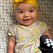 Emory joined the competition — help win amazing prizes! baby, smiling, yellow_headband, floral_dress, zigzag_blanket, infant, cute, happy, portrait, child, closeup, face, eyes, cheeks, hand, pattern, soft_lighting, indoors, toy, playful