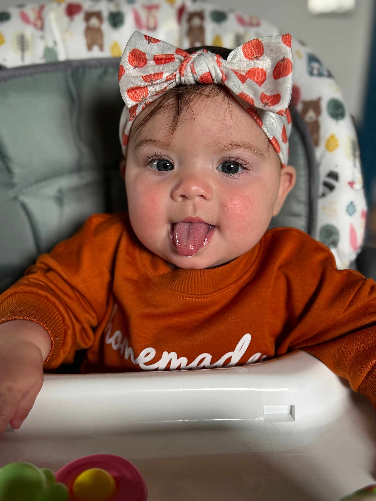 Emory is registered to the contest to win money with this photo: baby, child, high_chair, headband, tongue_out, orange_sweater, face, closeup, indoor, cute, infant, playful, expression, person, young_child, cheeks, clothing, seat, toy, background