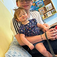 Pais is registered to the contest to win money with this photo: baby, teenager, sibling, hat, glasses, bookshelf, books, indoor, pillow, floral_shirt, smirk, holding, bracelet, shoe, denim, candid, portrait, child, family, seat