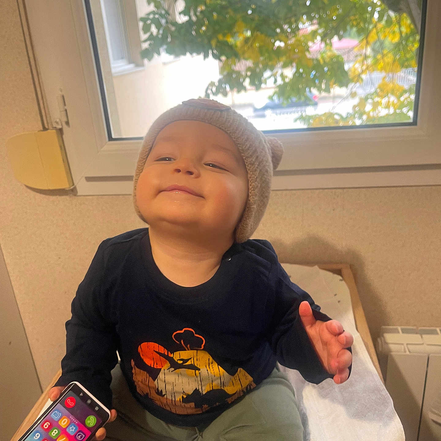 Gabriel participe au concours pour gagner de l'argent avec cette photo : baby, child, cozy, cushion, face, happy, hat, indoor, knit_hat, leaf, long_sleeve_shirt, pants, person, play, seated, smartphone_toy, smile, toy, tree, window
