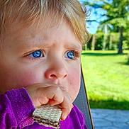 Sélène participe au concours pour gagner de l'argent avec cette photo : toddler, child, blue_eyes, purple_shirt, wafer, snack, outdoor, greenery, face, hand, close_up, young_child, food, nature, daylight, curly_hair, expression, sitting, garden, portrait