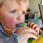 Sélène a rejoint le concours — aidez-le/la à gagner de superbes lots ! blonde_hair, blue_eyes, blurred_background, casual_clothing, child, close_up, daylight, drinking, expression, face, focus, hand, outdoor, person, portrait, pouch, rosy_cheeks, skin, toddler, young_child