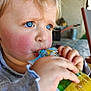 toddler, child, blue_eyes, blonde_hair, rosy_cheeks, drinking, pouch, close_up, portrait, hand, face, outdoor, blurred_background, casual_clothing, expression, young_child, skin, person, daylight, focus